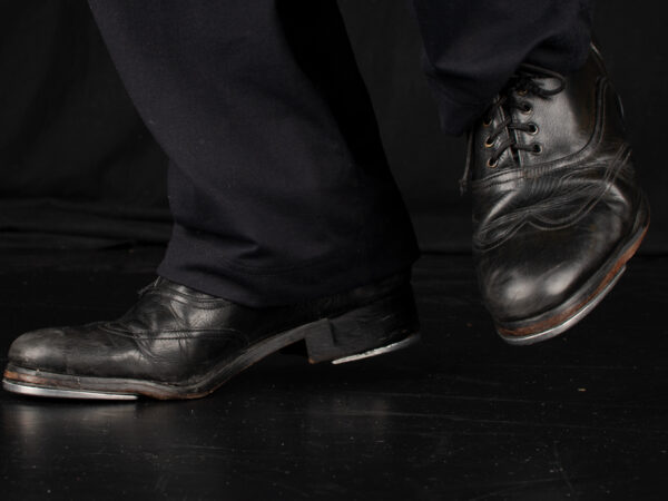 Eine Person in schwarzer Kleidung und schwarzen Stepptanz-Schuhen mit Schnürsenkeln auf einem dunklen Boden, wobei die Füße und Beine im Fokus stehen.