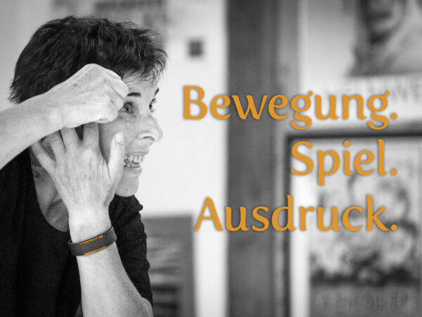 (Foto: Netty Kinsky) Schwarz-Weiß-Fotografie einer Pantomimin, die sich mit konzentriertem Gesichtsausdruck für ihren Auftritt schminkt. Ihre Hand berührt ihr Gesicht, während sie in den Spiegel blickt. Der Hintergrund zeigt unscharf einen Spiegel und ein gerahmtes Bild, was eine ruhige, vorbereitende Stimmung vermittelt. Das Bild betont die Kunst des körperlichen Ausdrucks und die Hingabe zur Pantomime.