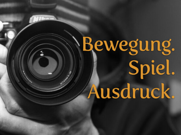 Schwarz-Weiß-Fotografie einer Kamera mit großem Objektiv, das von zwei Händen gehalten wird. Der orangefarbene Schriftzug „Bewegung. Spiel. Ausdruck.“ hebt sich deutlich ab und symbolisiert die zentralen Themen des Fotowettbewerbs. Das Bild betont die Verbindung von technischer Präzision und künstlerischem Ausdruck, die für die Einreichungen des Wettbewerbs stehen.