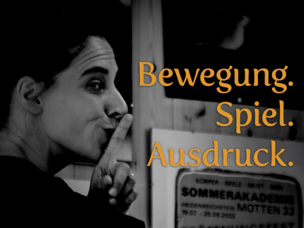 Schwarz-Weiß-Porträt von Nina Hlava, deren Finger nachdenklich an den Lippen ruhen, während der orangefarbene Schriftzug „Bewegung. Spiel. Ausdruck.“ die künstlerische Philosophie des Festivals betont. Im Hintergrund ist ein unscharfes Schild mit dem Hinweis auf eine Sommerakademie zu erkennen. Das Bild vermittelt die Intensität und den persönlichen Ausdruck, der im Mittelpunkt der darstellenden Künste steht. Foto: Netty Kinsky.