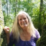 Samadhi Coulibaly Foto von Samadhi Coulibaly, die lächelnd in einer natürlichen, bewaldeten Umgebung steht. Sie trägt ein blau-lila gemustertes Oberteil und verkörpert die Freude und Offenheit, die ihre Biodanza-Workshops bei Waldviertel Exklusiv auszeichnen.
