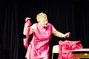 Irmgard Koller in einem leuchtend pinken Clownin-Kostüm: Sie hält eine pinkfarbene Handtasche in die Höhe, während ihr Gesicht und ihre Geste die Freude und den Witz ihrer Darstellung auf der Bühne des Festivals einfangen. Der dunkle Hintergrund betont die lebendige Präsenz und den künstlerischen Ausdruck. (Foto: C. Inel)