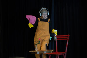 Sieglinde Svatos in orangefarbenem Overall und gelben Handschuhen auf der Bühne des Pantomime-, Clownerie- & Stepptanz-Festivals „Bewegung. Spiel. Ausdruck.“. Sie hält einen pinken Putzlappen in der einen und eine Sprühflasche in der anderen Hand, neben ihr ein Tisch mit einer Glasvase und einer roten Rose sowie einem Flachmann aus Edelstahl. Der dunkle Hintergrund unterstreicht die ausdrucksstarke Pose und die künstlerische Atmosphäre. (Foto: C. Inel)