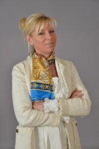 Barbara Stadler, Moderatorin des Pantomime-, Clownerie- & Stepptanz-Festivals „Bewegung. Spiel. Ausdruck.“, steht selbstbewusst vor einem einfarbigen Hintergrund. Sie trägt eine helle, strukturierte Jacke mit einem farbenfrohen Schal und hat die Arme verschränkt. Ihr Lächeln vermittelt Offenheit und Verbundenheit. (Foto: Barbara Stadler/Archiv)