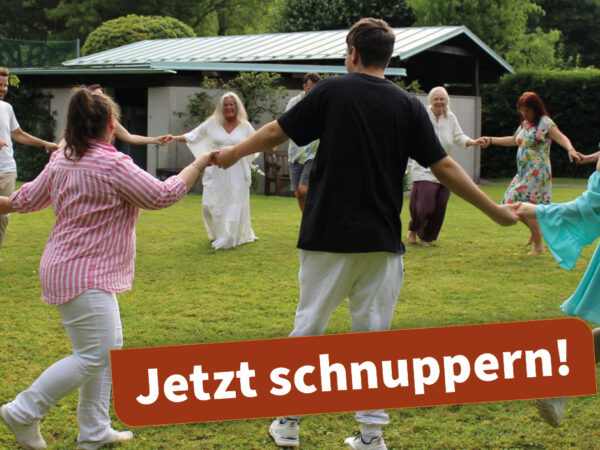 Foto eines Biodanza-Workshops im Freien: Eine Gruppe von Menschen tanzt im Kreis auf einer Wiese, umgeben von Bäumen und Bergen. Die Teilnehmer:innen sind in lockere, farbenfrohe Kleidung gekleidet und wirken entspannt und verbunden. Im Hintergrund ist ein kleines Gebäude zu sehen.