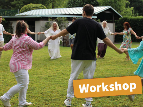 Foto eines Biodanza-Workshops im Freien: Eine Gruppe von Menschen tanzt im Kreis auf einer Wiese, umgeben von Bäumen und Bergen. Die Teilnehmer:innen sind in lockere, farbenfrohe Kleidung gekleidet und wirken entspannt und verbunden. Im Hintergrund ist ein kleines Gebäude zu sehen.
