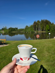 Foto einer Tasse Kaffee in einer Hand über einer grünen Wiese mit einem ruhigen See und Wald im Hintergrund. Aufnahme während der 2. Waldviertler Haustiermesse „Hund & Co“ im Hanfdorf Reingers – ein Moment der Entspannung inmitten der Natur. (Foto: © Sonja Wiesmüller - Vivenso)