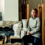 Portraitfoto von Lydia Reichberg: Die Malerin und Kunstpädagogin sitzt im Atelier zwischen ihren Werken – einem Landschaftsbild, einer Skulptur und einer Vase.